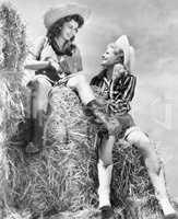 Two women in cowboy hats sitting on a haystack