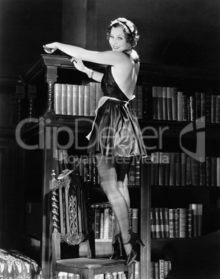 Portrait of a young woman standing on a chair and dusting a bookshelf in a sexy outfit