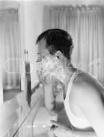 Profile of a young man in front of a mirror in a bathroom shaving