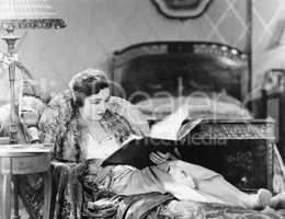 Young woman reclining in an armchair and reading a book in her bed room