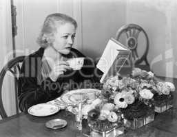Young woman sitting at the dining table holding a cup of tea and reading a book