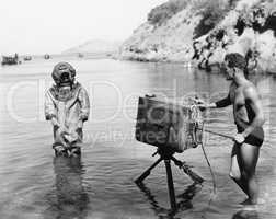 Profile of a young man holding a camera with a scuba diver standing in front of him on the beach