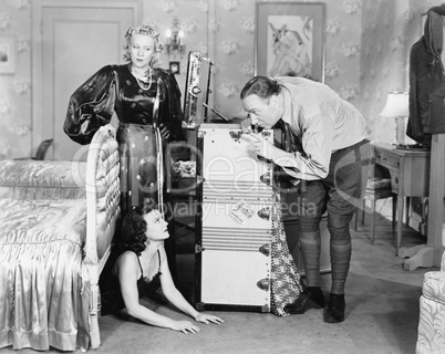 Young woman hiding under a bed and a couple standing near her