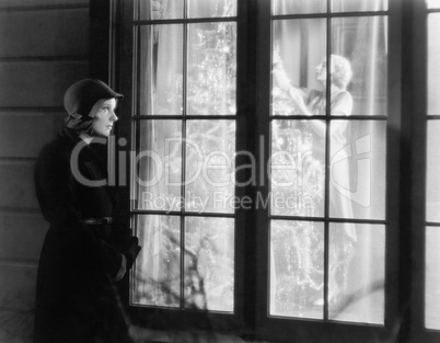 Woman standing outside of a window watching a woman trimming a Christmas tree