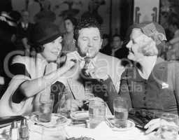Two women lighting a cigarette for a man