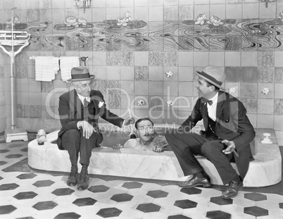 Two men sitting at the edge of a bathtub in suits and scrubbing a friends back