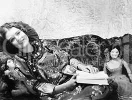 Woman in a Chinese print dress lying on the sofa with her dolls and a book