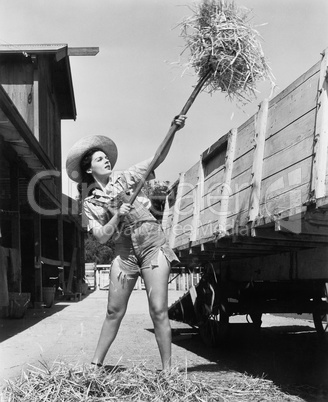 Young woman wearing cut off jeans and working at the farm pitching hay into a wagon