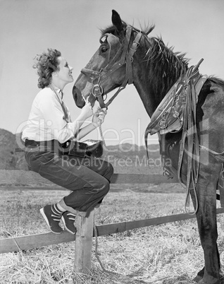 Young woman sitting on a fence and feeding her horse