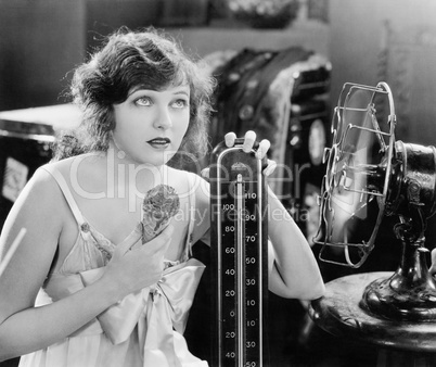 Young woman sitting next to a fan and a thermometer looking hot and eating an ice cream