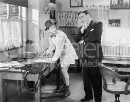 Two men standing in an office, one ironing his pants