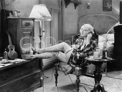 Young woman in her bedroom sitting on a chair with her legs on a dresser and a table with a few bottles of alcohol next to her