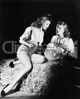 Two women sitting on a bale of hay carving a pumpkin