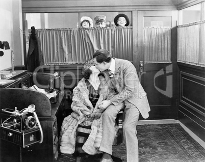 Couple kissing in an office while children and a messenger are peeking over a curtain