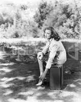 Young woman sitting on the side of the road on her suitcase, rubbing her aching feet