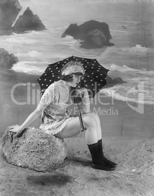 Young woman sitting on a bolder on the beach, holding an umbrella