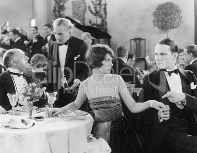 Young woman holding a man's hand at a different table while her companion is talking to the waiter