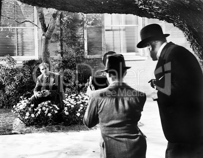 Men taking a picture of a man sitting in flower bed