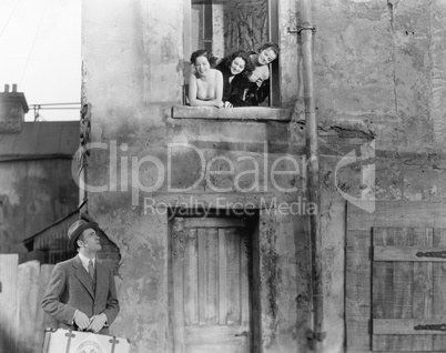 Three women looking out of a window at a man standing in the street with a suitcase