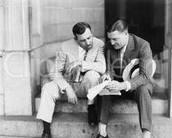 Two men sitting on steps and reading a document