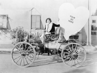 Young woman sitting on a car holding a big heart shaped sign