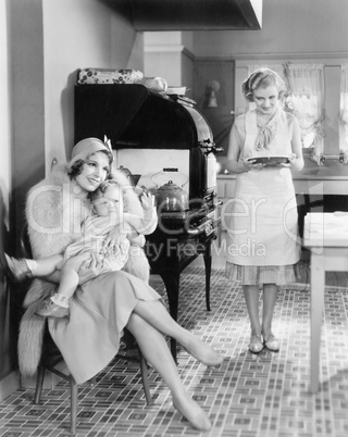 Elegant woman sitting with her baby in a kitchen while a friend is bringing a pie