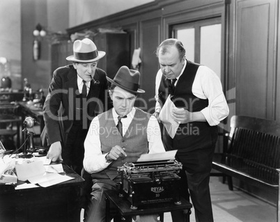 Three men in an office hunched over a typewriter