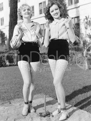 Two young women standing together playing horse shoes