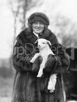 Woman in a fur coat and hat holding a small baby lamb in her arms
