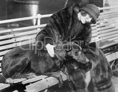 Woman in a fur coat sitting on a bench petting her dog