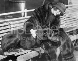 Woman in a fur coat sitting on a bench petting her dog