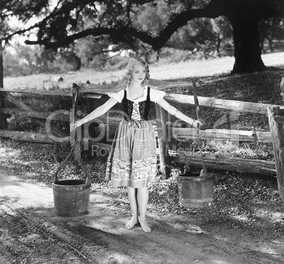 Barefoot woman in a tied bodice dress carrying water buckets on shoulder poles
