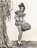 Woman in short polka dot dress in front of cornstalk carrying pumpkin