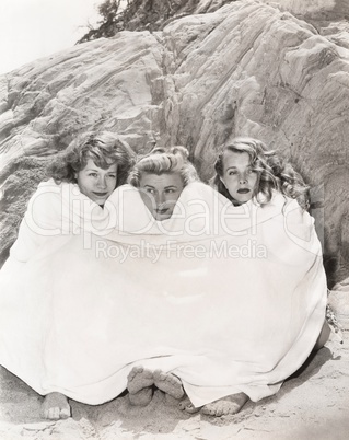 Three women huddled under a towel on beach