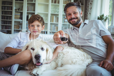 Father and son sitting on sofa with pet dog and watching television