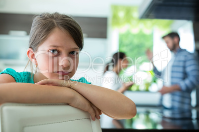 Sad girl leaning on chair while parents arguing in background