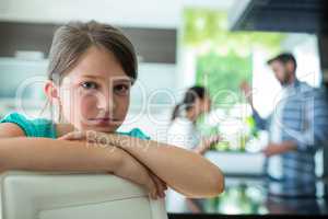 Sad girl leaning on chair while parents arguing in background