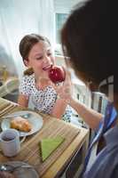 Mother feeding apple to her daughter while having breakfast