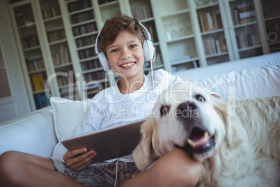 Boy sitting on sofa with pet dog and listening to music on digital tablet
