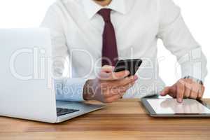 Mid-section of businessman using mobile phone and digital tablet with laptop on table