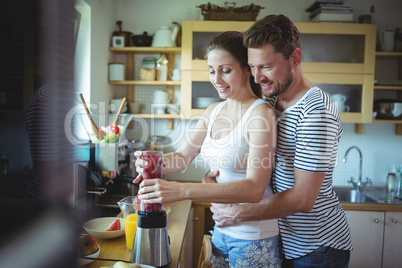 Man embracing woman from behind while preparing watermelon smoothie in the kitchen