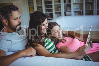 Parents lying on sofa with daughter and using digital tablet in living room