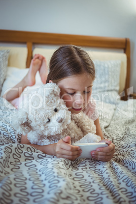 Happy girl lying on bed with a teddy bear and using mobile phone