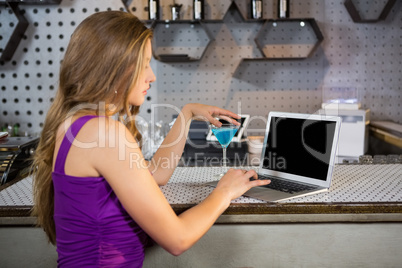 Beautiful woman using laptop while having cocktail