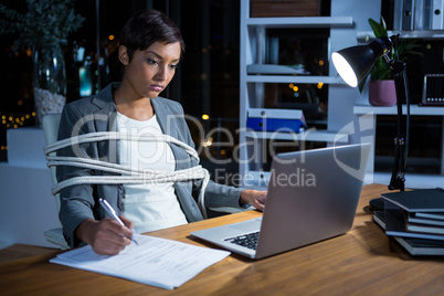 Businesswoman tied with rope while working on laptop at her desk