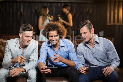 Three happy friends looking at mobile phone while having cigar and whisky