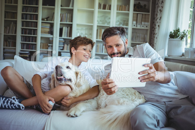 Father and son sitting on sofa with pet dog and using digital tablet
