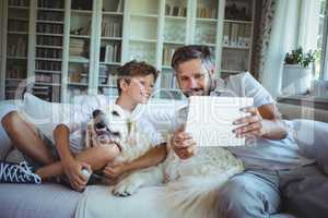 Father and son sitting on sofa with pet dog and using digital tablet