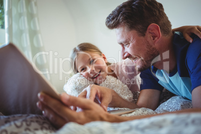 Father lying with daughter on bed and using digital tablet