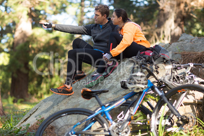 Biker couple sitting on rock pointing at distance in forest